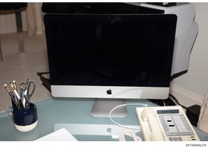 This photograph shows a contemporary office workspace featuring an Apple iMac desktop computer, wireless mouse, office telephone system, desk organizer with writing implements, and other standard office equipment on a light blue desk surface. The ima