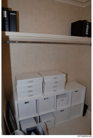 This is an evidence photograph depicting an interior storage room or closet space containing multiple white filing boxes and storage containers organized on shelves and the floor. The room features beige/tan walls, white crown molding, and what appea