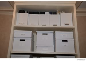 This photograph documents an organized storage shelving unit containing multiple white document boxes and file containers, each labeled with category tabs. The boxes are systematically arranged on two shelves and appear to contain organized records, 