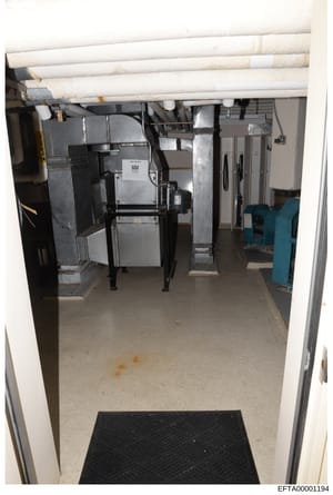 This is a photograph of a basement or utility room showing HVAC systems, ductwork, and various industrial or mechanical equipment. The room contains heating/cooling units, metal ductwork overhead, electrical panels, and blue machinery visible in the 
