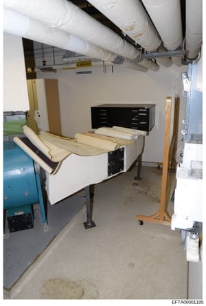 This evidence photograph shows a basement or utility room space containing HVAC ductwork, a black filing cabinet or storage unit, rolled materials (possibly blueprints or architectural documents), industrial equipment including a blue cylindrical uni