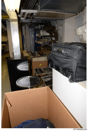 This photograph shows a storage or evidence room containing various seized items including cardboard boxes, luggage, electronic equipment, and other materials. The image appears to be part of the evidence documentation from the investigation, showing