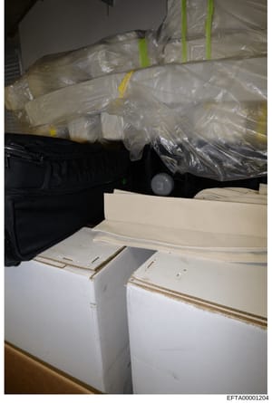 This is an evidence photograph showing multiple sealed plastic-wrapped packages with yellow and green evidence tape markers, stacked alongside white cardboard boxes. The image appears to be from a property seizure or evidence collection operation, wi