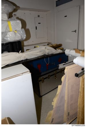 This is a photograph of an evidence storage or property room containing seized items wrapped in protective plastic and stored on shelving. The image shows a blue/black trunk or container in the center, large plastic-wrapped bundles (some marked with 