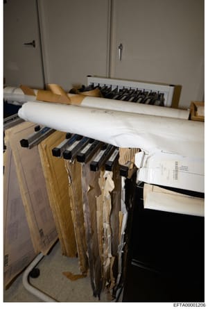 This photograph shows organized evidence storage in what appears to be an FBI evidence room or secure facility. Multiple documents are systematically hung from horizontal bars using tan and cream-colored folders with black clasps, with rolled bluepri