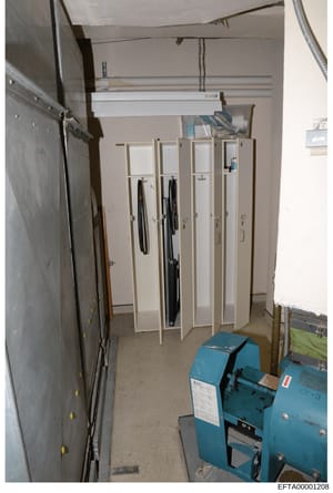 This is a photograph of an interior room containing four white storage lockers with clothing hanging inside, industrial equipment in blue (possibly a compressor or similar machinery), overhead storage racks, and various tools and cables. The room app
