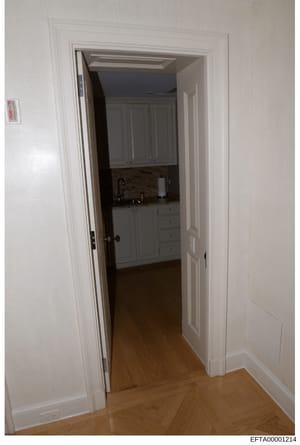 This is a documentary photograph showing the interior of a residential space, specifically a view through a doorway into a kitchen area. The image captures white trim work, wooden cabinetry, countertops, and hardwood flooring. This type of photograph