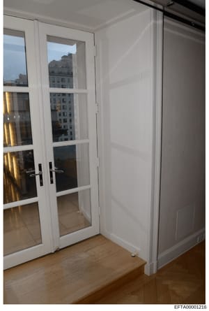 This photograph shows an interior room with white-framed glass double doors opening to what appears to be a balcony or terrace. Through the doors, a view of urban apartment buildings is visible. The room features white walls, wooden flooring, and mod