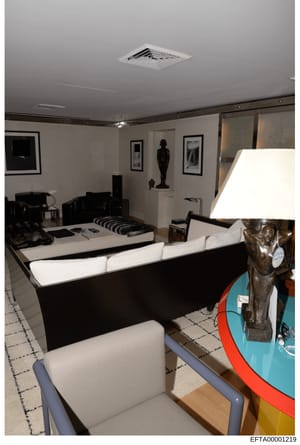 This photograph depicts the interior of a yacht cabin showing a spacious entertainment area with contemporary décor. The room features modern black and white furniture including a large sectional sofa, built-in bed with striped bedding, framed black 