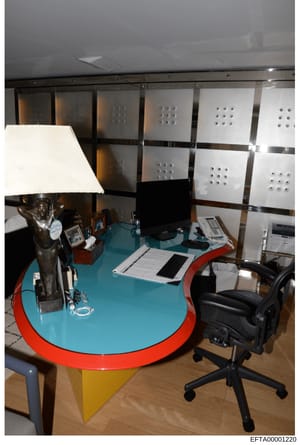 This is a photograph of a modern office interior featuring a distinctive curved turquoise desk with red trim and yellow support base. The space includes office equipment, storage cabinets, and furniture consistent with a professional workplace or res