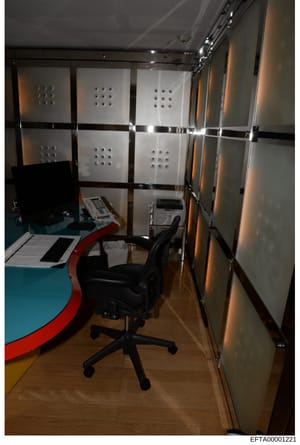 This photograph documents an interior space containing office equipment and storage infrastructure, likely taken as part of the evidence collection process during the investigation. The room features a desk workspace with a black office chair, organi