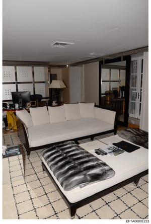 This photograph shows a residential living room or lounge area featuring contemporary furniture and décor. The space includes a white upholstered sofa, a bed or daybed with a dark patterned mattress cover, a desk workstation with computer equipment, 