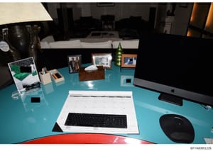 This photograph shows a turquoise desk setup in what appears to be a residential office or study space. The desk contains a computer monitor, keyboard, calendar (dated 2019), framed photographs, decorative items, and various office supplies. The imag