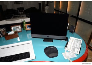 This photograph shows a workspace desk with a turquoise surface containing an Apple iMac computer, office equipment including a telephone and calendar, personal photographs, and various desk accessories. The image appears to be evidence documentation