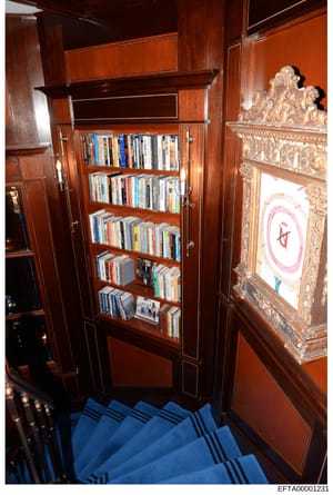 This is an evidence photograph from an Epstein property showing an interior hallway or room featuring a wooden bookcase with glass doors containing DVDs and books, along with ornate decorative artwork and framing. The image documents the interior fur