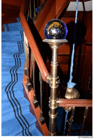 This is an evidence photograph showing the interior of a property, featuring a wooden staircase with blue carpeting and a decorative brass stair post topped with a blue lapis lazuli sphere adorned with gold leaf. The photo documents the upscale finis