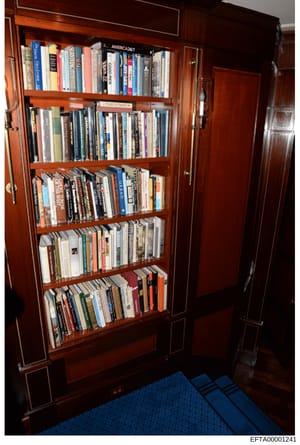 This is an evidence photograph showing a wooden bookshelf with glass doors located in what appears to be a residential interior space. The shelf contains a large collection of books, DVDs, and other media materials organized across six shelves. The p
