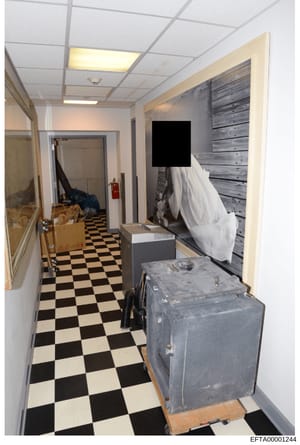 This is a photograph of an interior basement or utility hallway showing storage items, equipment, and general conditions. The image depicts a space with distinctive black and white checkered flooring, drop ceiling with recessed lighting, and various 