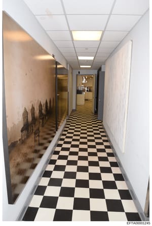 This photograph shows a long, narrow hallway with a black and white checkered tile floor, fluorescent ceiling panels, and white walls. On the left wall is artwork depicting what appears to be a cityscape or architectural silhouette. The hallway exten