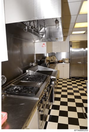 This is a photograph of a professional or institutional kitchen space, featuring commercial-grade cooking equipment including a large gas range/stove, hood ventilation system, and stainless steel countertops. The kitchen appears to be part of a resid