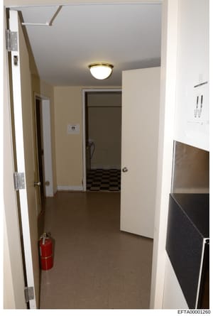 This is an evidence photograph showing an interior hallway with cream-colored walls, a ceiling fixture, and multiple doors. The space appears to be part of a building under investigation, with a red fire extinguisher visible on the floor. The photogr