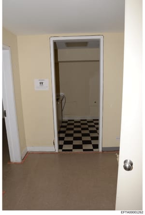 This is an evidence photograph showing the interior of a room with pale yellow walls, a doorway with white trim leading to a space with black and white checkered flooring, and a visible light switch marked 'VV'. The photo appears to be part of proper