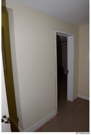 This is a photograph of an interior hallway or corridor space showing cream-colored walls, a white-framed doorway, and dark carpeting with red/orange trim along the baseboard. The image appears to be evidence documentation from a property search or i