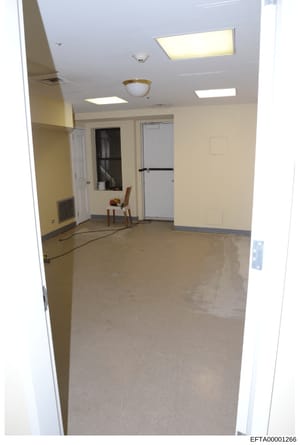 This is a photograph of an empty, sparsely furnished interior room with institutional or commercial characteristics. The room features pale yellow walls, recessed ceiling lights, exposed HVAC vents, and minimal furnishings including a single wooden c