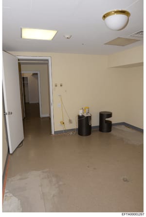 This photograph shows an empty, unfinished interior room with concrete flooring, beige walls, and white ceiling with lighting fixtures. The space appears to be under construction or renovation, with exposed electrical wiring, two black cylindrical co
