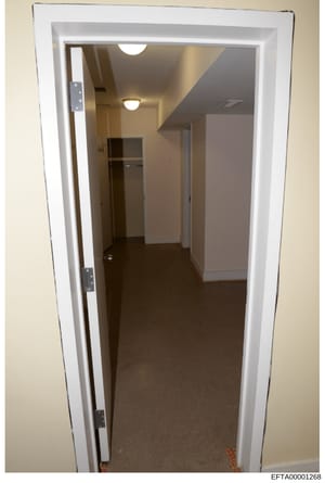This is a photograph of an interior hallway or corridor, likely taken as part of property documentation or evidence collection during the investigation. The image shows a narrow passage with white-trimmed doorways, fluorescent ceiling lighting, dark 