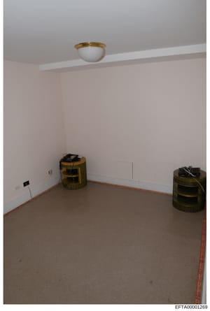 This is an evidence photograph showing an empty interior room during renovation or investigation. The room contains two woven cylindrical storage containers with black coverings and wiring, a ceiling-mounted light fixture with brass trim, and appears