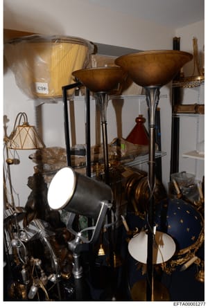 This photograph documents the contents of a storage or display room containing various lighting fixtures, decorative home furnishings, and accessories. The image shows floor lamps with bronze bowl shades, spotlights, woven baskets, ornamental spheres