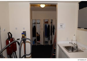 This is an evidence photograph showing the interior of a utility or service room, featuring a large closet with glass doors containing hanging clothing and personal items, cleaning equipment stored on the left side, and a sink on the right. The photo