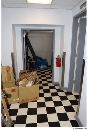 This is an evidence photograph showing an interior storage or utility room with a distinctive black and white checkered floor. The room contains multiple cardboard boxes, blue material/bags, and a red fire extinguisher mounted on the wall. The photog
