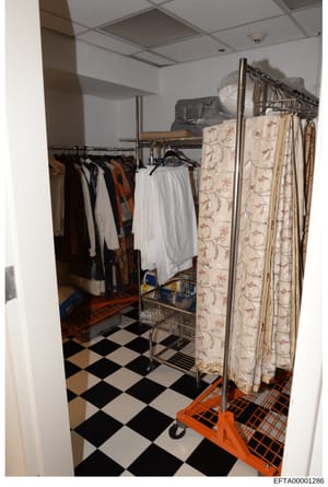 Evidence photograph showing an interior storage space or closet area with clothing racks, assorted garments, decorative panels, and household items. The image documents the contents and condition of a room, likely from an Epstein-related property. Th