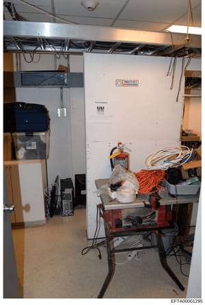 This is an investigative photograph showing the interior of a basement storage room containing various electrical equipment, tools, and materials. The room features exposed ceiling infrastructure, electrical panels, fire safety equipment, coiled cabl