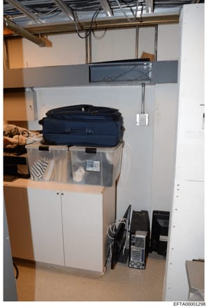 Evidence photograph showing a storage or utility space containing a dark blue suitcase on a clear plastic storage bin, white cabinet storage below, and computer equipment on the floor. This appears to be documentary evidence from a property search or