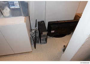 Evidence photograph showing a storage or utility room containing discarded or stored electronic equipment including computer towers, monitors, a humidifier, and other household items. The image appears to be from a property search or seizure related 