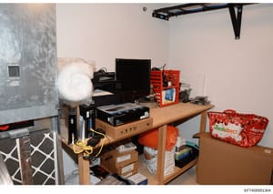 This is an evidence photograph showing an interior room containing computer equipment, networking hardware, storage boxes, and household items. The image appears to be from a search or seizure operation, documented with a Bates number for investigati