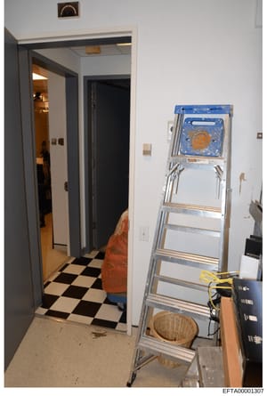 A photograph showing an interior room or hallway with a black and white checkered floor, an open doorway leading to another space, and a metal ladder positioned against the right wall. Various household items and electrical equipment are visible. The