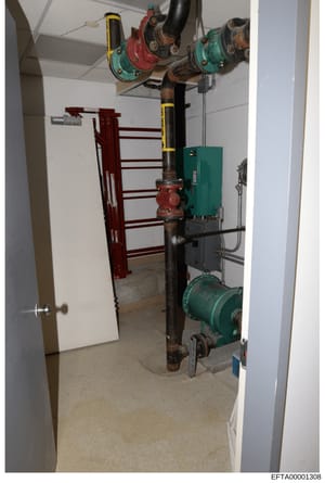 This is a photograph of a basement or utility room showing mechanical and HVAC equipment including pulleys, pipes, pumps, and a heating/cooling unit. The image documents the building's infrastructure and may be part of property inventory or structura