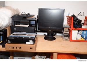 This is an evidence photograph showing seized computer equipment and networking devices displayed on a wooden desk. The image includes an HP multifunction printer, a TRENDnet network switch in a cardboard box, a computer monitor, and various cables a