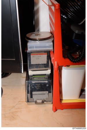 This is an evidence photograph showing a stacked storage device containing multiple media items and materials. The device bears a handwritten label reading '#17 JEE call' and is positioned next to a red storage rack. The image documents the physical 