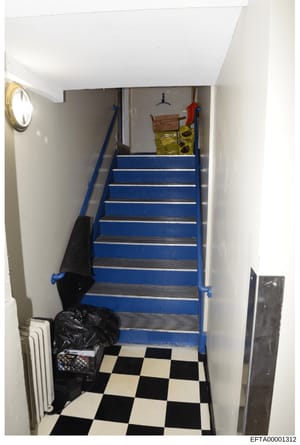 This is a photograph of an interior stairwell, likely from a residential or commercial property under investigation. The image shows a flight of blue and metallic steps leading upward, with white walls, a checkered black and white floor, and various 