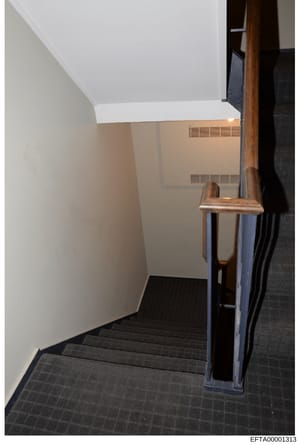 This photograph shows an interior stairwell with dark tile flooring, white/beige painted walls, a wooden stair railing, and visible HVAC vents on the upper walls. The image appears to be documentation of a residential or commercial interior space, li