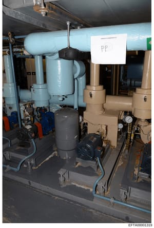 This is a photograph of industrial mechanical equipment, likely part of a building's HVAC or plumbing system. The image shows blue and tan-colored pipes, pumps with motors, gauges, and a black valve or connector. A white label marked 'PP' is visible 