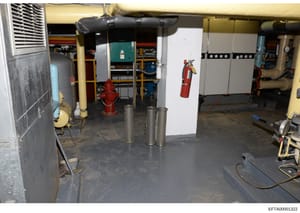 This photograph shows an interior mechanical or utility room containing building systems infrastructure, including HVAC equipment, piping, fire suppression systems, and safety equipment. The image documents the basement or service area of a building 