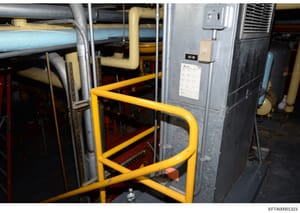 This photograph documents the interior mechanical systems of what appears to be a large residential or commercial property, showing HVAC equipment, piping, and control systems. The image captures yellow safety railings, air conditioning units, plumbi