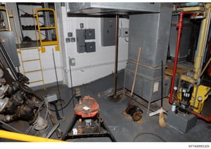 This is a photograph of an industrial or maintenance workspace containing various mechanical equipment, electrical panels, hydraulic systems, and tools. The image shows a basement or ground-level utility room with multiple large machines, electrical 