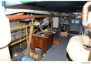 This is a photograph of a mechanical room or basement area containing HVAC equipment, piping systems, and various building infrastructure. The image shows industrial equipment including an orange pipe, yellow safety railings, copper piping, electrica